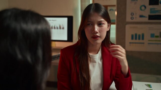 Two young Asian businesswomen arguing about a financial report during a late-night meeting in the office, showing signs of stress, disagreement, and conflict over project results