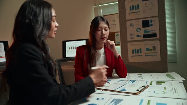 Young Asian businesswomen engaging in a serious discussion while analyzing financial charts and graphs on documents during a late-night meeting in a modern office. Working overtime to meet a deadline
