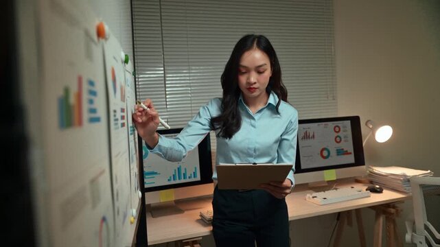 Dedicated Asian businesswoman working late in the office, standing at a whiteboard and analyzing financial charts and graphs with a pen while holding a clipboard with important documents