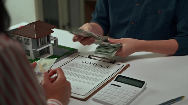 Professional realtor counting a large stack of dollar bills from a client as a down payment for a new home purchase, with a contract, calculator, and house model on the office desk