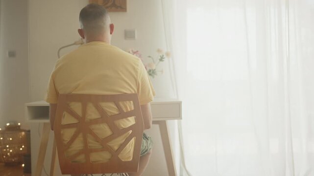Rear view of an overweight or obese man with a buzz cut sitting on a modern wooden chair at a desk, contemplating and looking out the window of his sunlit apartment during the day