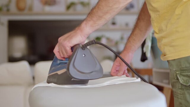 Slow motion of a man's hands ironing a white garment on an ironing board in a living room, with steam coming out of the hot electric appliance. Domestic chores and daily household routine concept