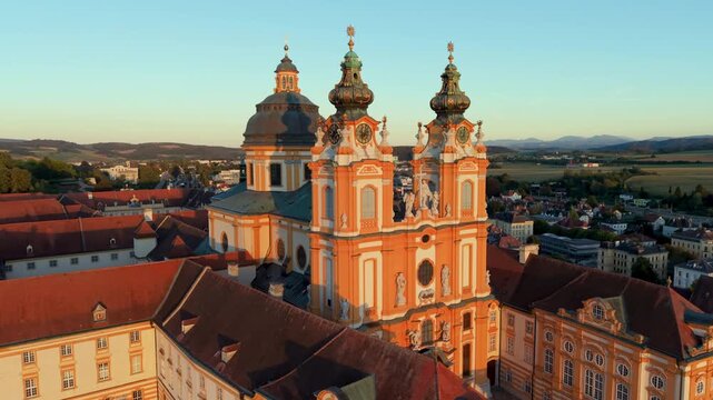 Aerial panorama of baroque monastery on hill during golden hour showing warming sunlight and lengthening shadows across terracotta roofs and riverside landscape. Melk. Austria