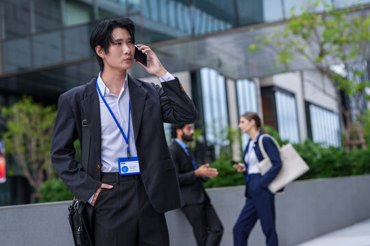 A focused young Asian executive pauses to check the screen of his mobile phone in front of luxury retail frontage in a central business district, colleagues blur softly in the background