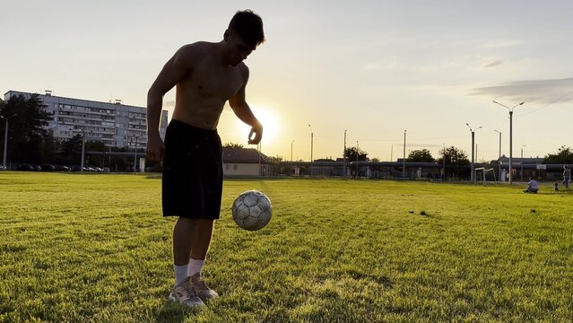 Young man juggling soccer ball on stadium at sunset. Professional footballer kicking ball at green field. Sportsman practicing tricks at meadow with sunlight at background. Freestyle football