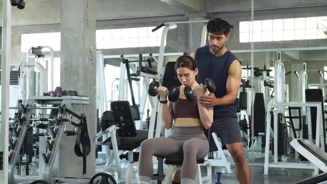 Personal trainer coaching a young woman during a dumbbell workout in a modern gym, promoting strength training, fitness, healthy lifestyle and professional exercise guidance.
