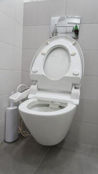 Modern toilet with bidet seat. Wall-mounted white toilet in tiled bathroom, electronic bidet seat with control panel on the left and lid raised, grey floor tiles and compact sanitary design, no people