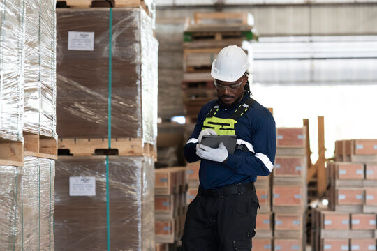 African American male warehouse worker wearing safety uniform, hard hat using technology wireless digital tablet checks stock inventory of plywood in industrial wood manufacturing factory warehouse