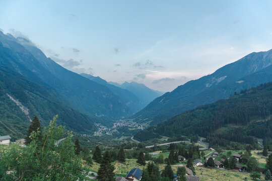Elevated panoramic view over a deep Swiss Alpine valley with scattered houses, green meadows, evergreen forest and winding mountain roads. Scenic rural landscape ideal for travel, tourism, countryside