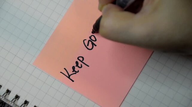 Female hand writes down Keep Going with black pen on a pink sticky note. Communication, message, reaching out, motivation, and encouragement.