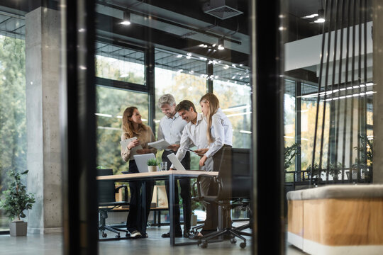 Group of business people standing together and discussing their work and projects, having a team meeting in an office, view from glass wall