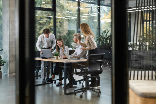 Group of business people standing together and discussing their work and projects, having a team meeting in an office, view from glass wall