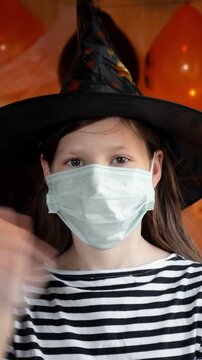 Vertical video of preteen girl in witch hat wearing medical face mask, kid looks at camera during halloween party celebration. Healthcare during seasonal holidays. Post-pandemic celebration concept