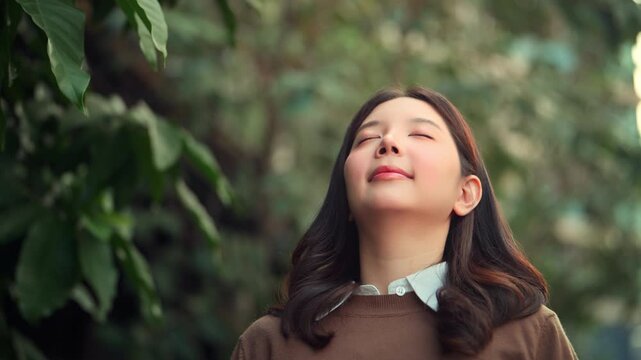 Young Asian woman closing her eyes and taking a deep breath in green urban park, peaceful female enjoying fresh air and quiet moment in nature, mindfulness, relaxation, wellness and stress relief conc