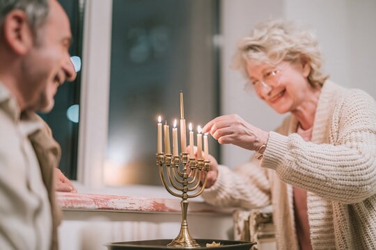 Brass menorah with glowing candles being lit by a senior woman and man during a reverent Hanukkah celebration for community and family