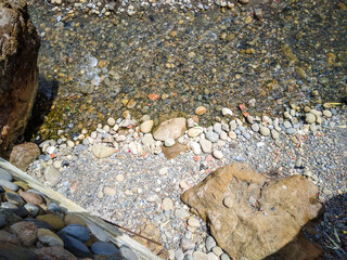 Crystal clear water flows gently over smooth multicolored river stones and pebbles near large natural rocks creating a beautiful texture in this bright sunny scene © Graphixa