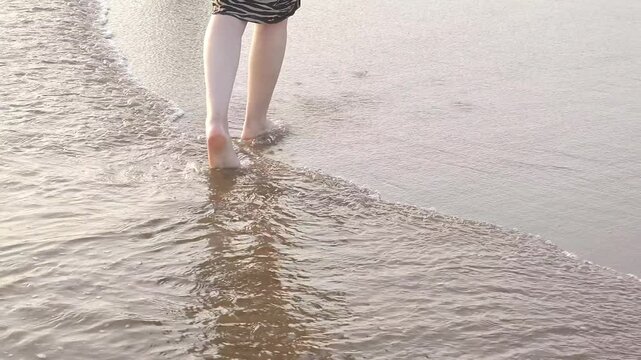 A woman in a long skirt is walking barefoot through the ocean waves, holding on to her shoes, and the water is splashing around her feet. Summer vacation, the concept of a beach holiday and relaxation