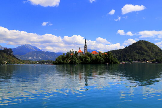 Lake Bled, Slovenia: Wide scenic view of the crystal clear turquoise lake water with tourist rowboats, the famous island with church, and mountains under a blue sky.