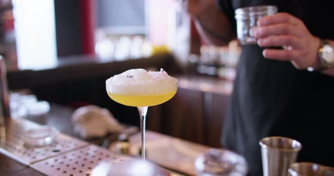 Decorating drink. Cocktail recipe. Bartender male hand tweezers putting dried flower petal to foam liquor preparing in coupe glass.