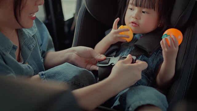 Asian mother fastening safety harness on toddler daughter sitting in child car seat, parent securing little girl before driving, childcare