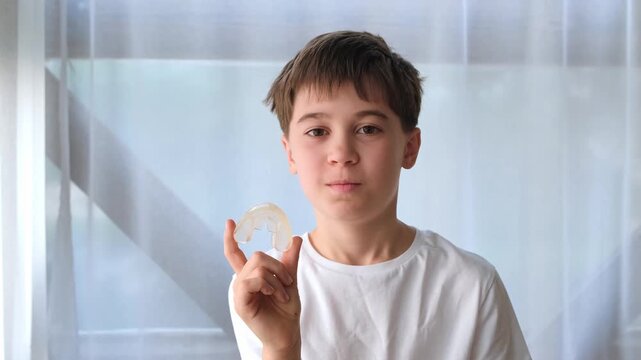 The smiling caucasian boy holds a dental aligner in his hand looking at camera. Orthodontic treatment, dental care, oral hygiene. Gap teeth or diastema. High quality 4k footage.