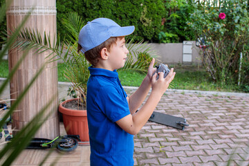 boy standing in yard holding joystick remote controller in both hands, potted plant and stone column, brick paved path, green hedge in background, one person, childhood leisure and technology © Татьяна Швец