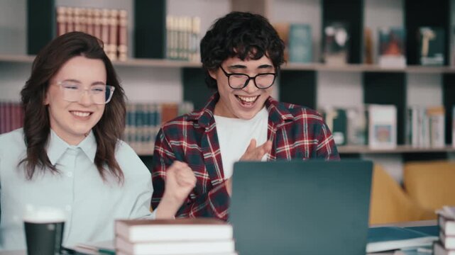 Students celebrating success on laptop with books and coffee in library study session