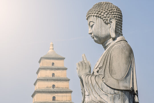 View of a stone Buddha statue in a prayer pose with the historic Giant Wild Goose Pagoda in the background under a clear sky in Xi'an, Shaanxi, China.