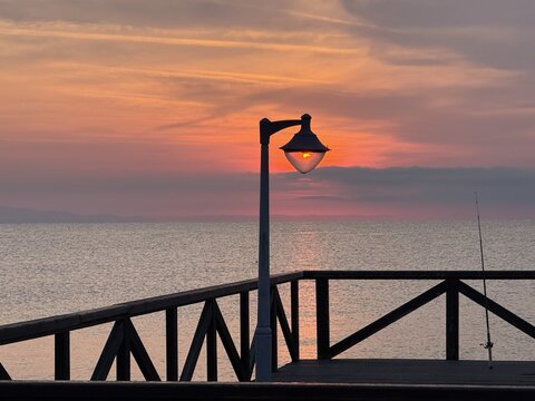 View of a sunset over the sea from a wooden pier with a lamp post and a fishing rod in Paralia, Katerini, Greece.