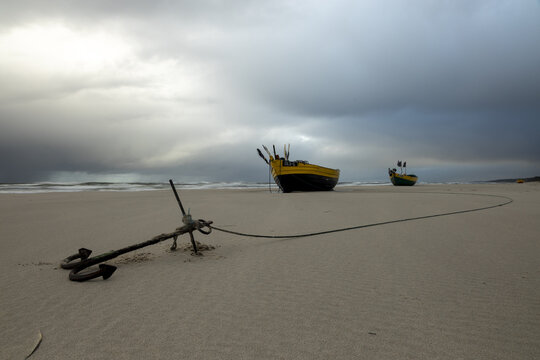 View of a rusty metal anchor on a sandy beach connected by a rope to a yellow and black fishing boat under a dramatic stormy sky in Poland.