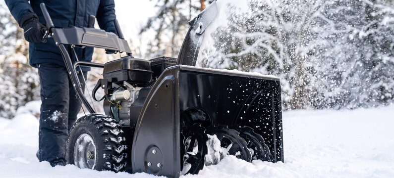 A person operates a snow blower to clear snow from a snowy yard. The scene shows trees covered in white snow, typical of a winter day
