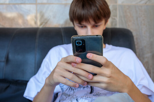 teen sitting on black leather sofa holding a smartphone with both hands, looking at screen, stone wall behind, one person, gadget addiction, smartphone dependence and youth problems concept