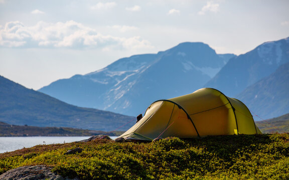 View of a yellow tent pitched on a grassy hill in Veslelangdalen, overlooking a lake with snow-capped mountains in the background in Fjord, More og Romsdal, Norway.