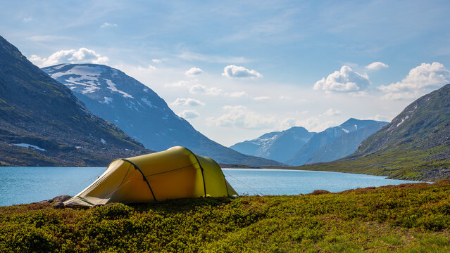 View of a yellow tent pitched on the grassy shore of a blue lake in the Veslelangdalen valley, surrounded by majestic mountains in Fjord, More og Romsdal, Norway.