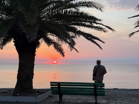 View of a man standing by a green bench under a palm tree watching a vibrant sunrise over the calm sea in Paralia, Katerini, Greece.