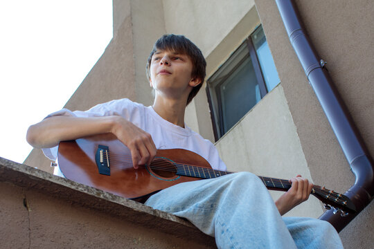 teenager sitting on rooftop ledge playing acoustic guitar, building wall with window and drain pipe behind, bright sky, low angle view, music hobby, leisure and free time outdoors in daylight