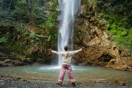 View of a man standing with arms outstretched in front of the cascading Tiger Falls surrounded by lush green forest and rocky cliffs in Dehradun, Uttarakhand, India.