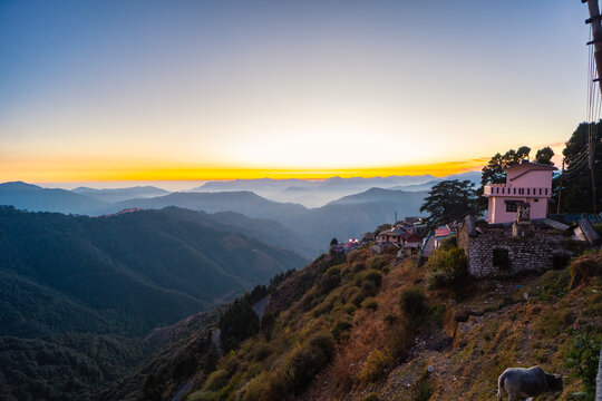 View of Chakrata village with stone buildings and a cow grazing on the hillside, overlooking the hazy Himalayan mountain range at sunset in Dehradun, Uttarakhand, India.