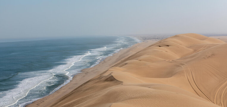 View of Skeleton Coast where massive sand dunes meet the Atlantic Ocean with tire tracks visible on the sand in Kunene Region, Namibia.