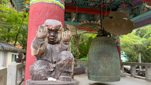 View of a traditional wooden statue of a child praying next to a large bronze Buddhist temple bell under a colorful pavilion roof at Jangsan Mountain, Busan, South Korea.
