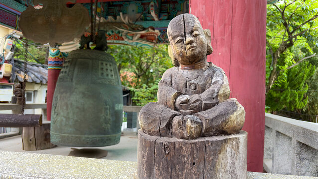 View of a weathered wooden monk statue sitting in meditation next to a large bronze temple bell in a traditional pavilion at Jangsan Mountain, Busan, South Korea.