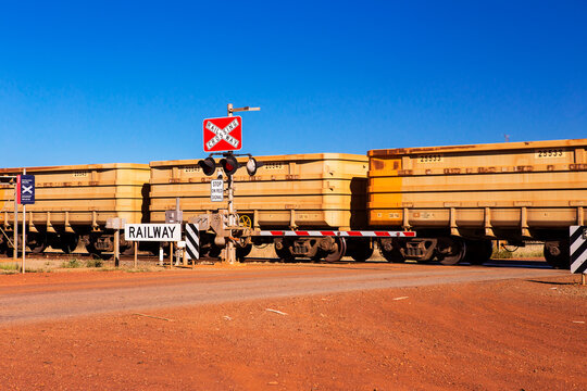 View of a long freight train passing through a railway crossing in the red desert landscape of the Pilbara region in Newman, Western Australia, Australia.