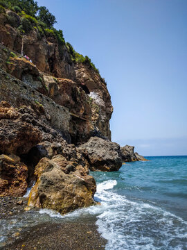 Massive rocky cliff face towers over the vibrant blue ocean waves splashing against the jagged shoreline on a bright and clear sunny summer afternoon day