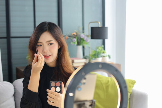 Beautiful Asian female beauty blogger live streaming makeup tutorial at home holding eyeshadow palette and brush in front of ring light.
