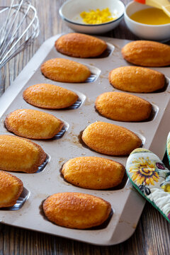 Freshly baked madeleines cooling in a metal baking tray on wooden table.