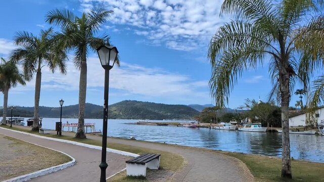 Slow motion video of the waterfront at Pontal Beach in Paraty, Brazil, featuring tall palm trees, colonial street lamps, stone benches, and tourist boats floating on the calm bay.