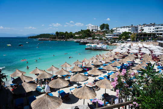 KSAMIL, ALBANIA - JULY 28, 2026: Sunny beach with turquoise water, straw umbrellas, and people relaxing, real lifestyle travel illustration.