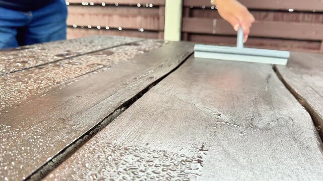 Close-up of a person using a rubber squeegee tool to wipe rain water droplets off an outdoor wooden patio table surface after rain.
