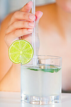 Woman is holding a clear glass filled with a light-colored drink, garnished with a lime slice and a straw.