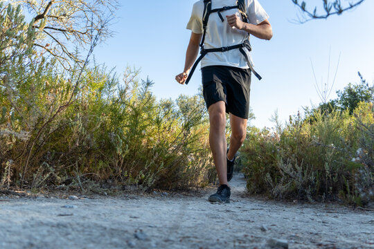 Man actively running on a dirt trail, wearing a backpack and athletic shorts, surrounded by nature and green bushes